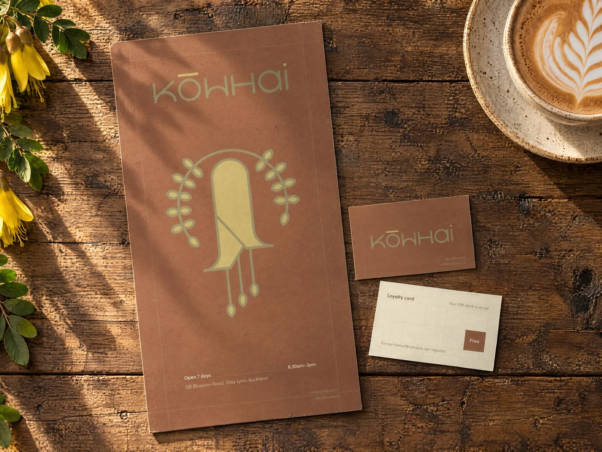 Overhead view of the Kōwhai menu card and loyalty card, front and back, on a weathered timber café table in warm morning light, beside a flat white and a sprig of yellow kōwhai flowers.